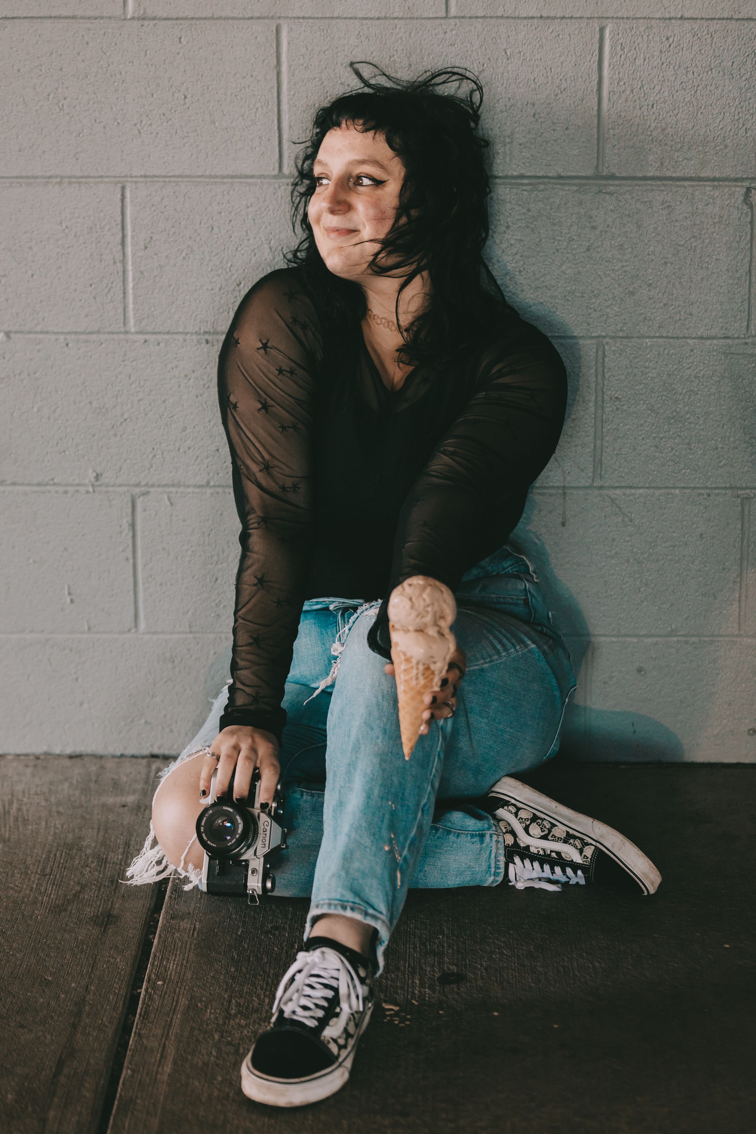 An image of a woman holding a camera and an ice cream cone sitting in front of a grey brick wall.