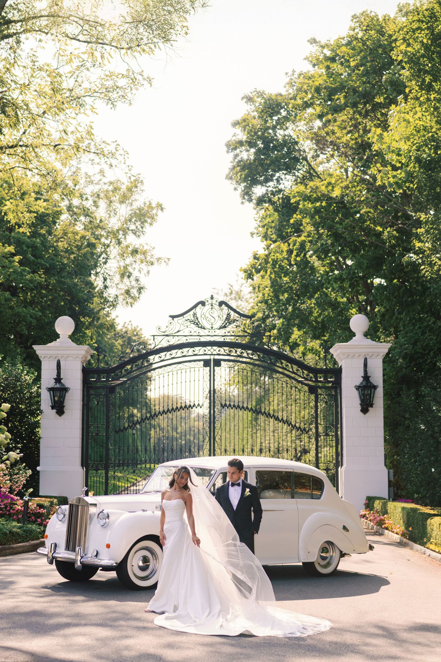 Bride and groom posing by a vintage white Rolls-Royce in front of elegant iron gates surrounded by lush greenery.