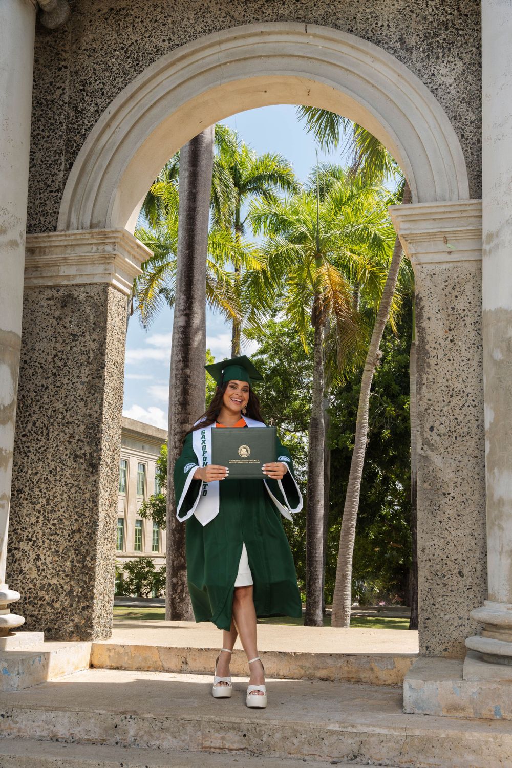 Graduate holds laptop while standing under classical stone arch with palm trees in background.