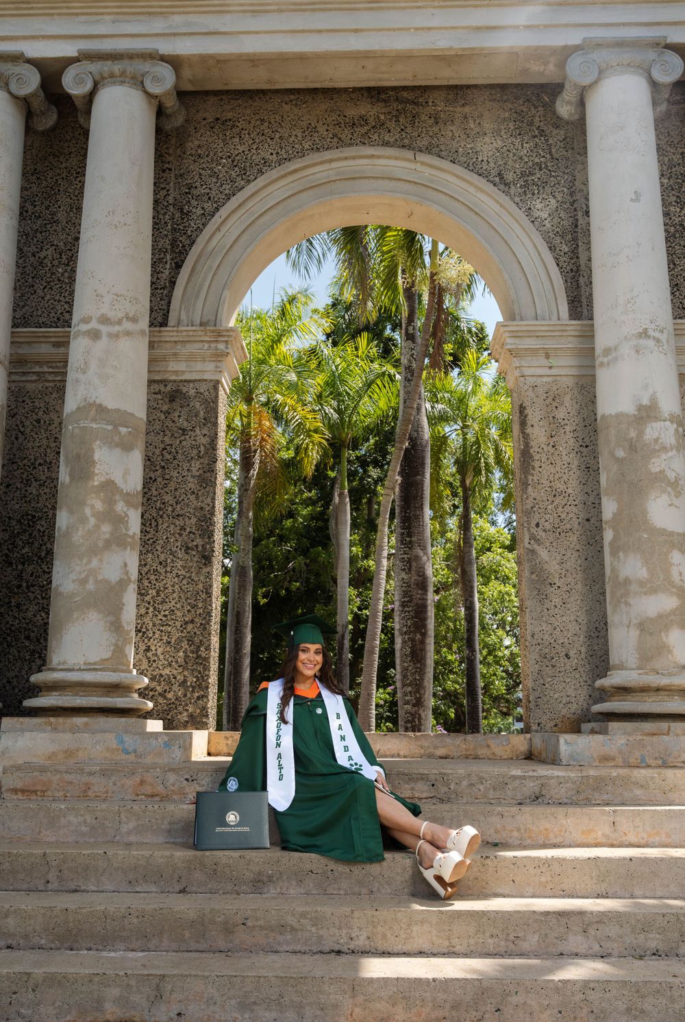 Graduate in green regalia poses under ornate stone archway with palm trees visible in background.