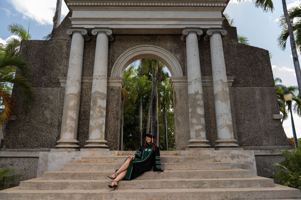 Wide view of stone archway with graduate sitting on steps surrounded by tropical landscaping.