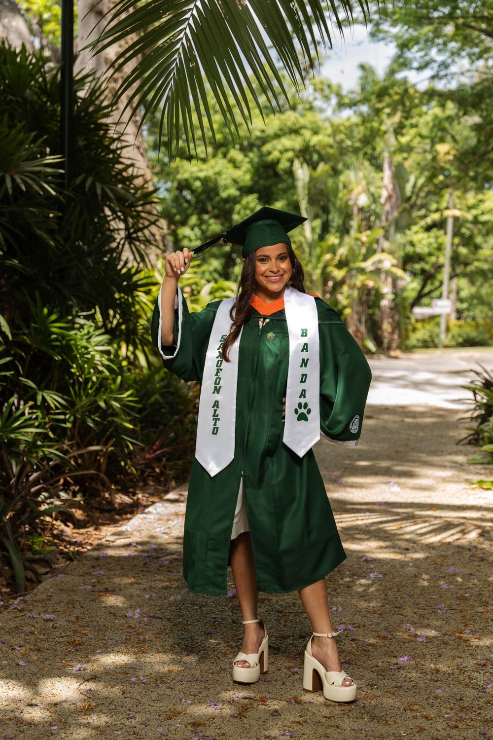 Graduate in green cap and gown poses with honors sash in tropical garden setting.
