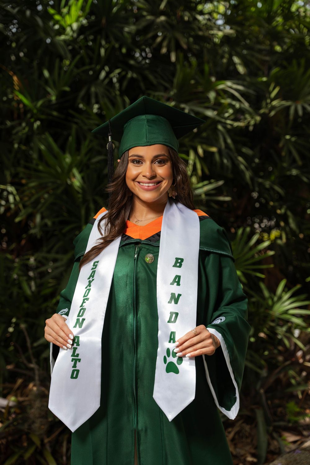 Graduate wearing green academic regalia displaying graduation stoles in tropical setting.