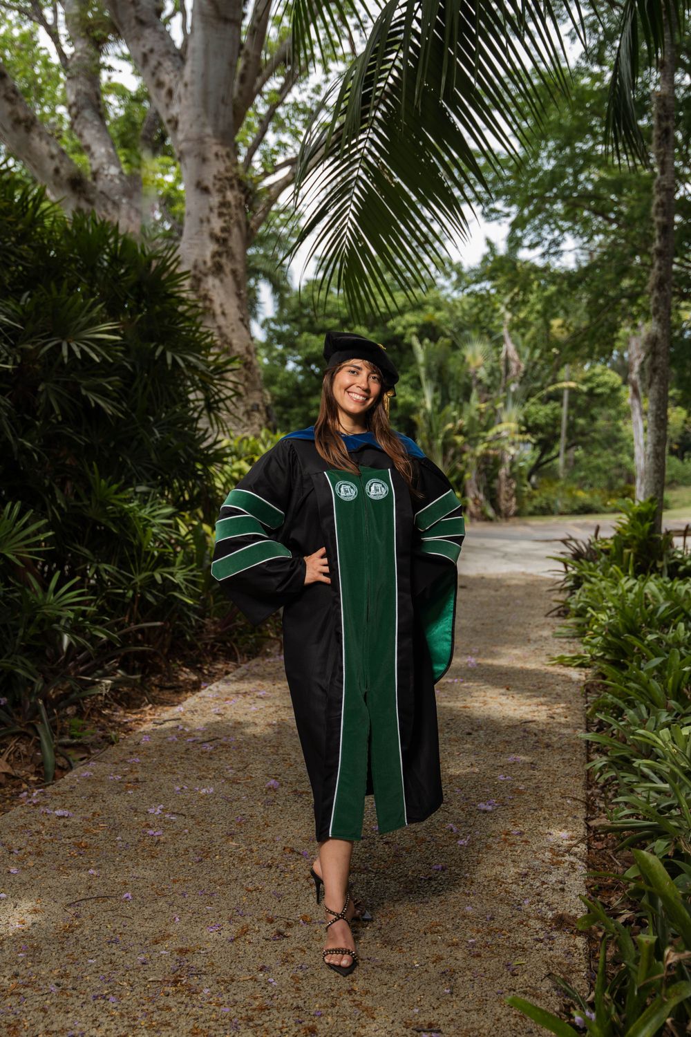 Graduate walking on garden path wearing dark green academic regalia.