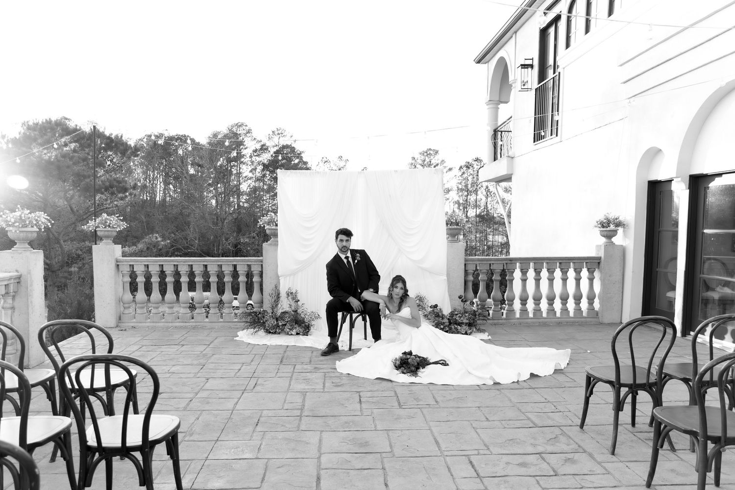 Black and white photo of a dramatic scene on an elegant stone terrace with vintage chairs and architectural details.