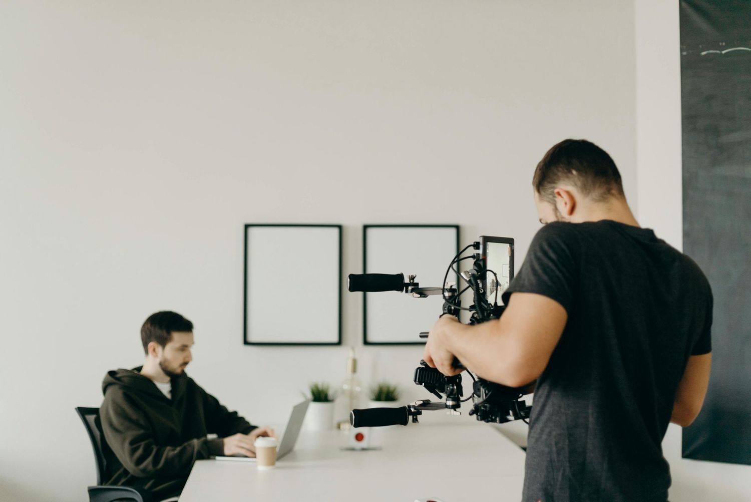 Professional videographer filming content creator working on laptop in minimalist office setting.