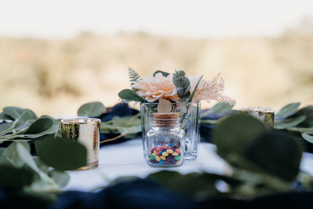 A decorative jar with flowers sits among green foliage on a wedding reception table.