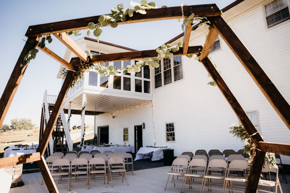 An outdoor wedding ceremony setup with wooden arch and white folding chairs against a white building.