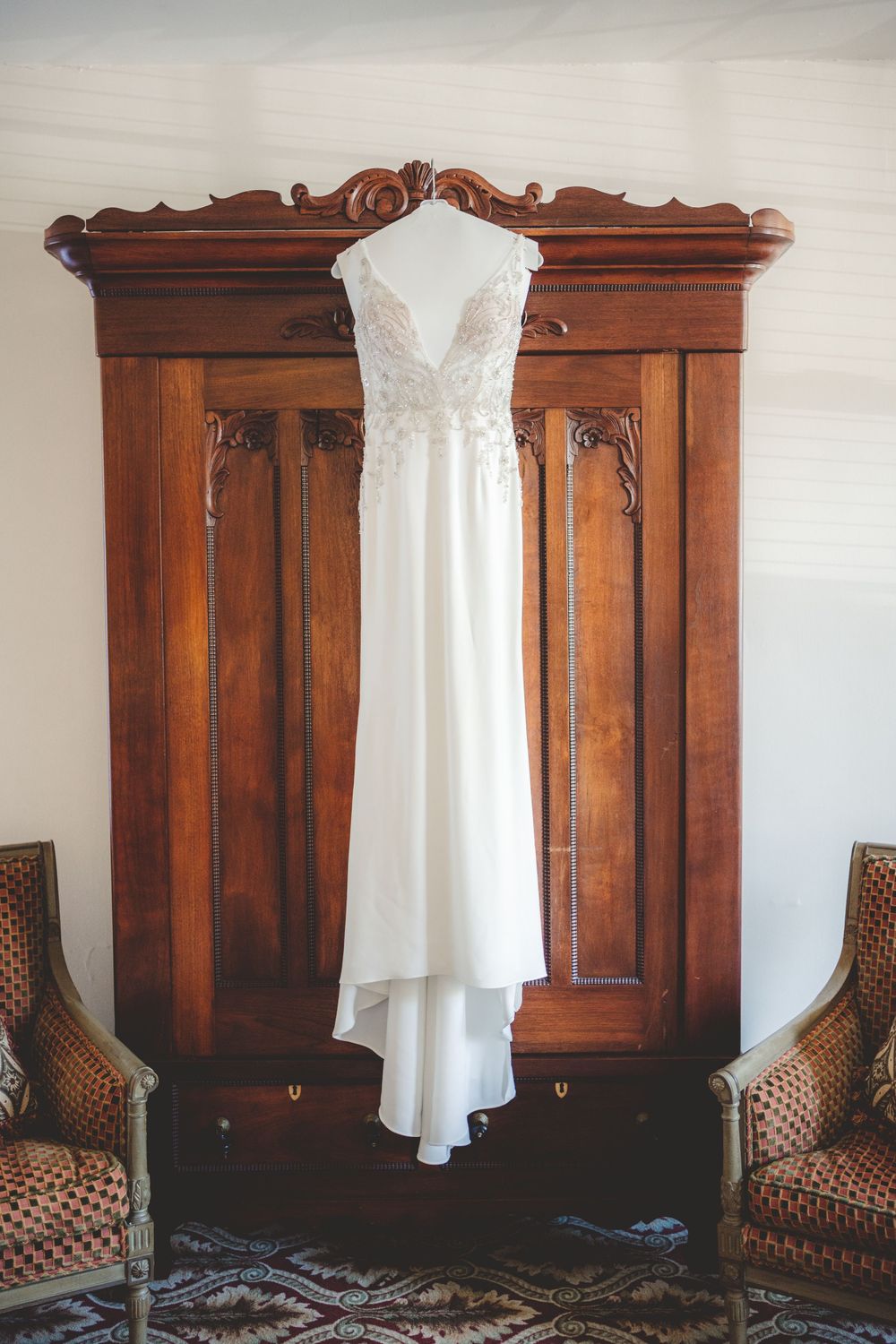 White wedding gown displayed on antique wooden armoire between two upholstered chairs.