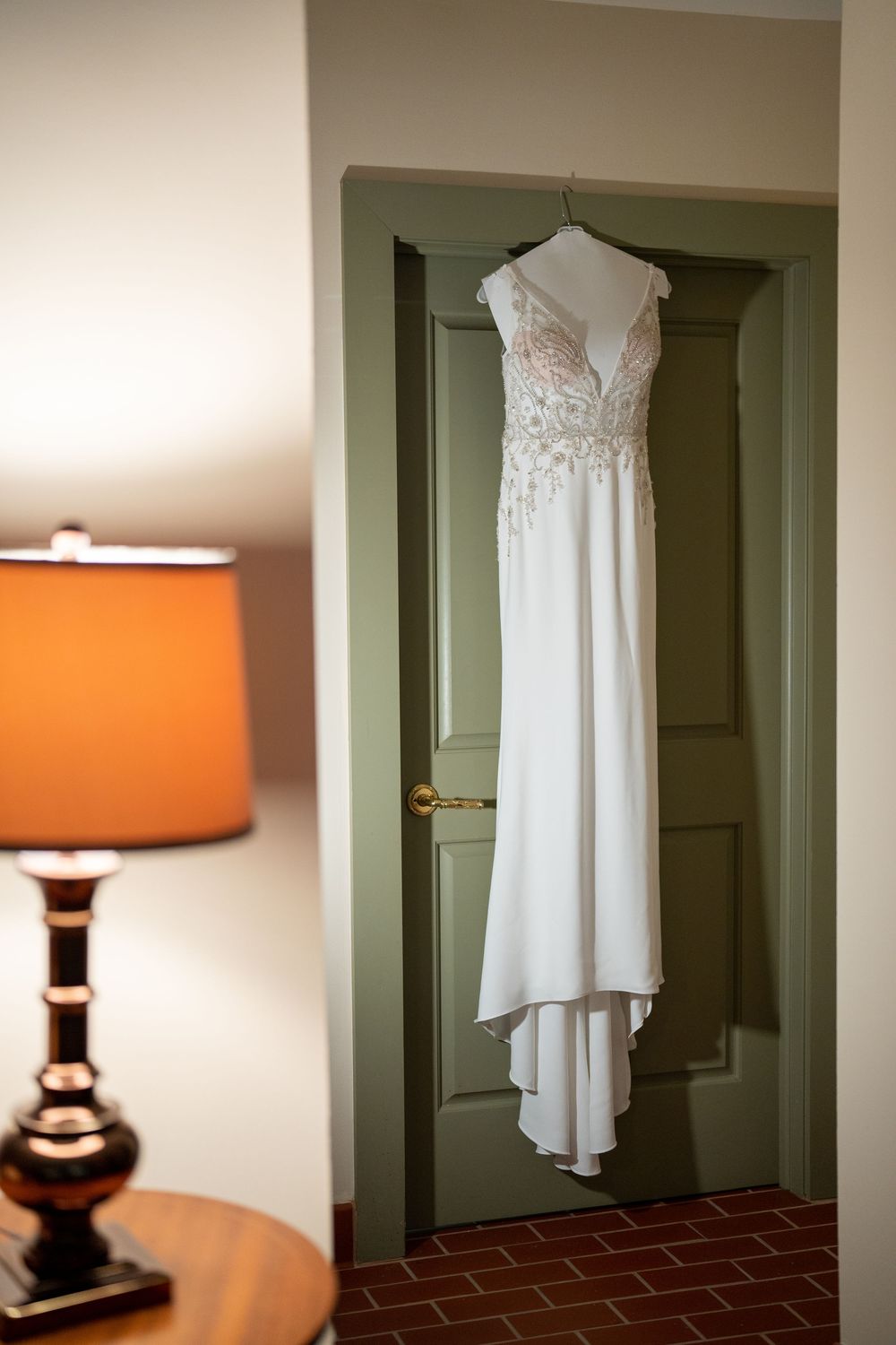 White wedding dress with lace bodice hanging on mint green door next to orange table lamp.