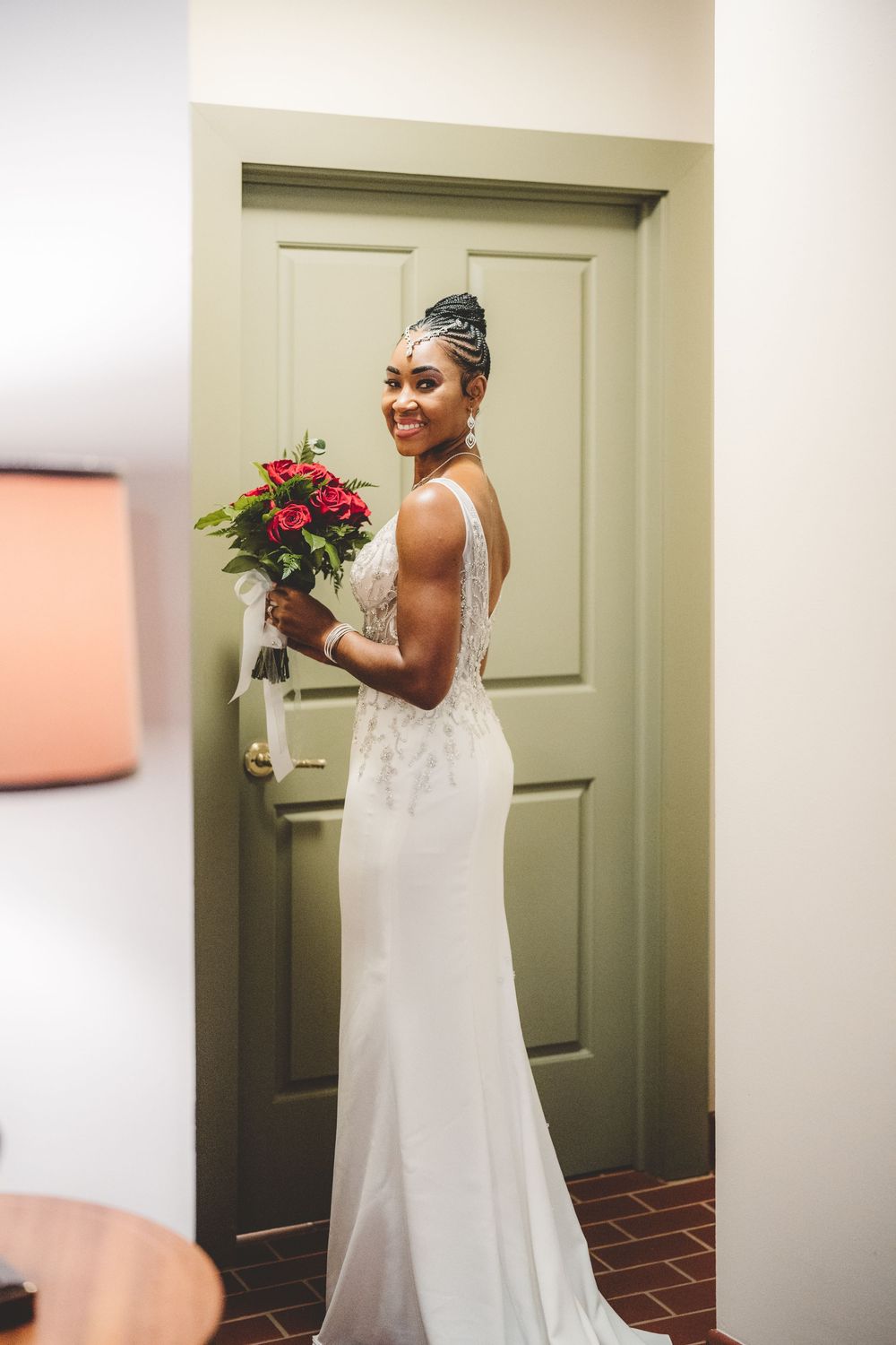 A bride in an elegant white dress holds a red bouquet while standing in front of a sage green door.