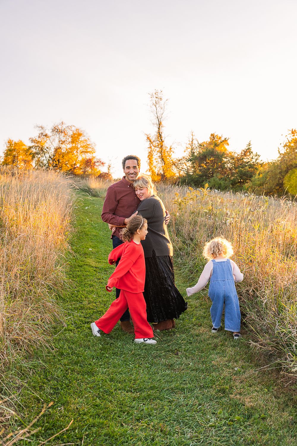 A family walks together on a path through tall autumn grass at sunset.