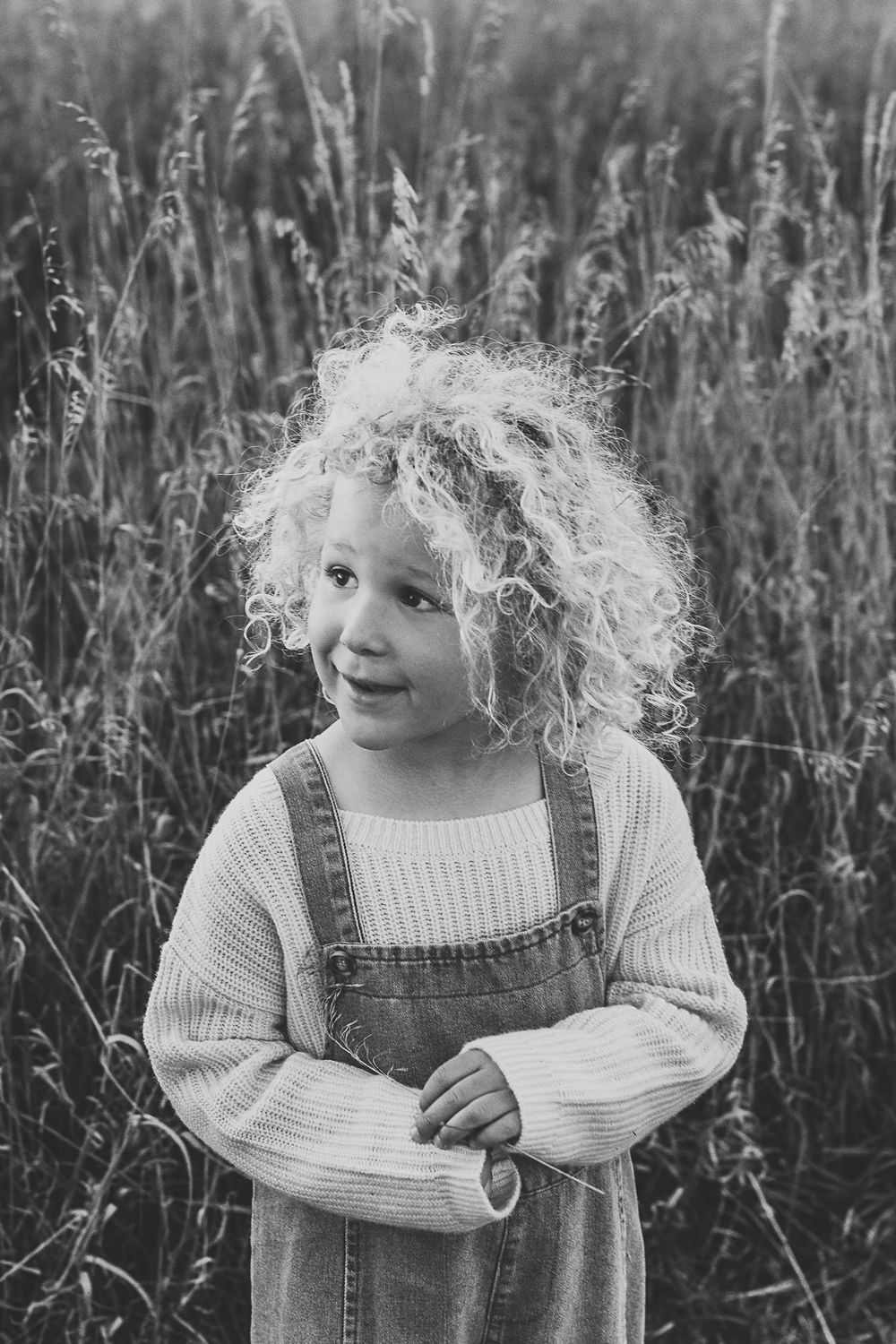 A black and white portrait of a child with curly hair wearing overalls and a sweater.