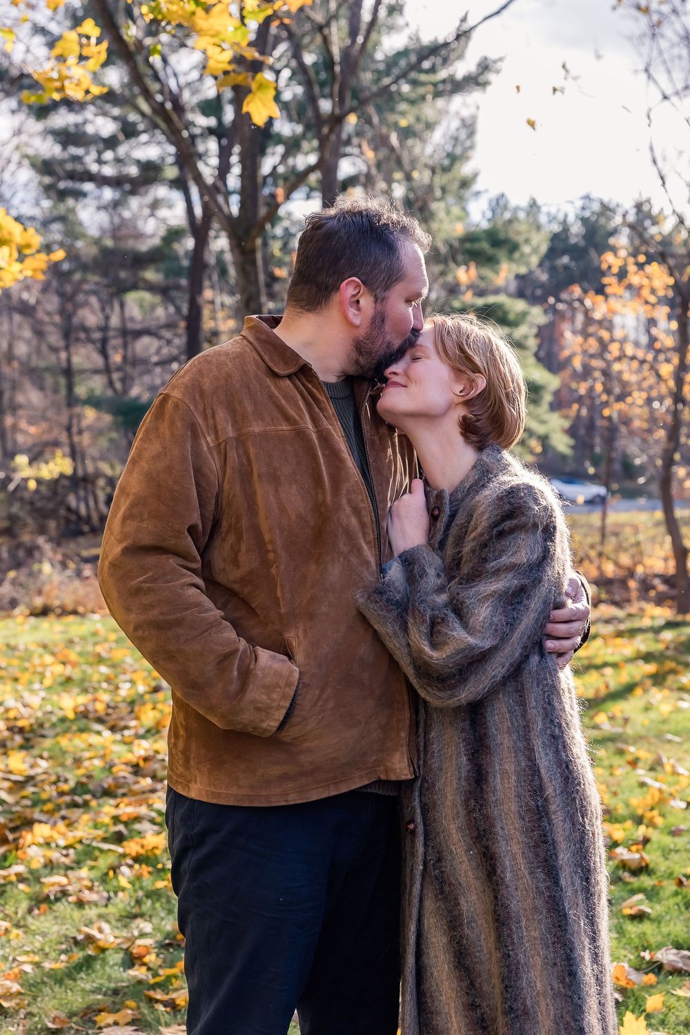 A couple shares an intimate kiss outdoors in autumn with golden leaves and pine trees in the background.