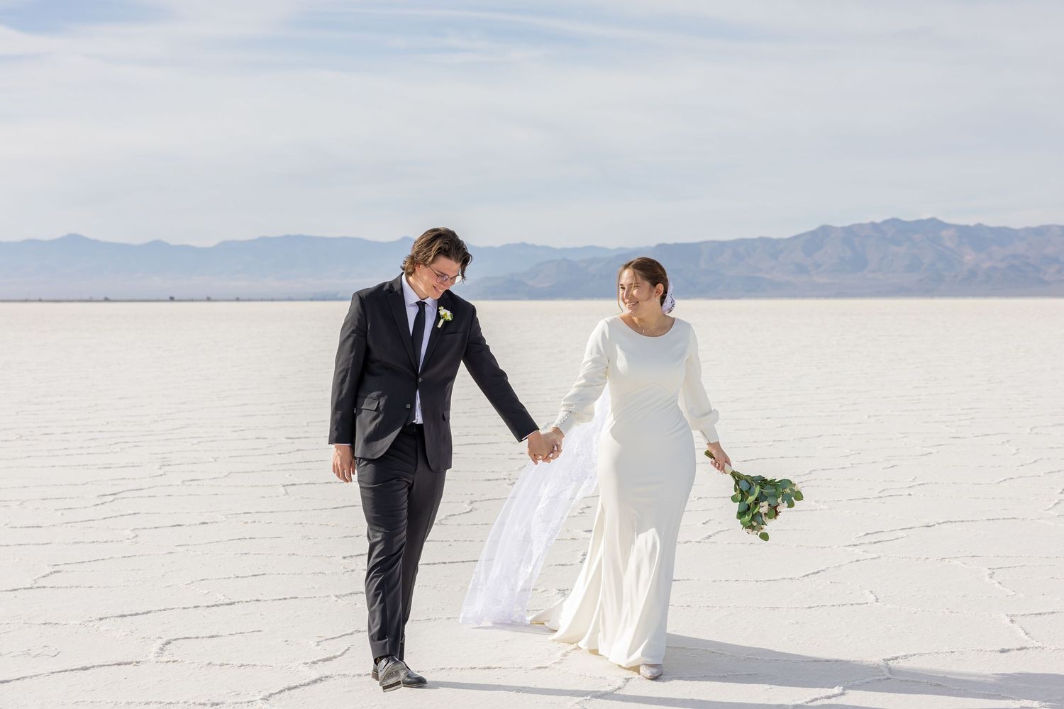 Couple walking together on white salt flats under bright blue sky.