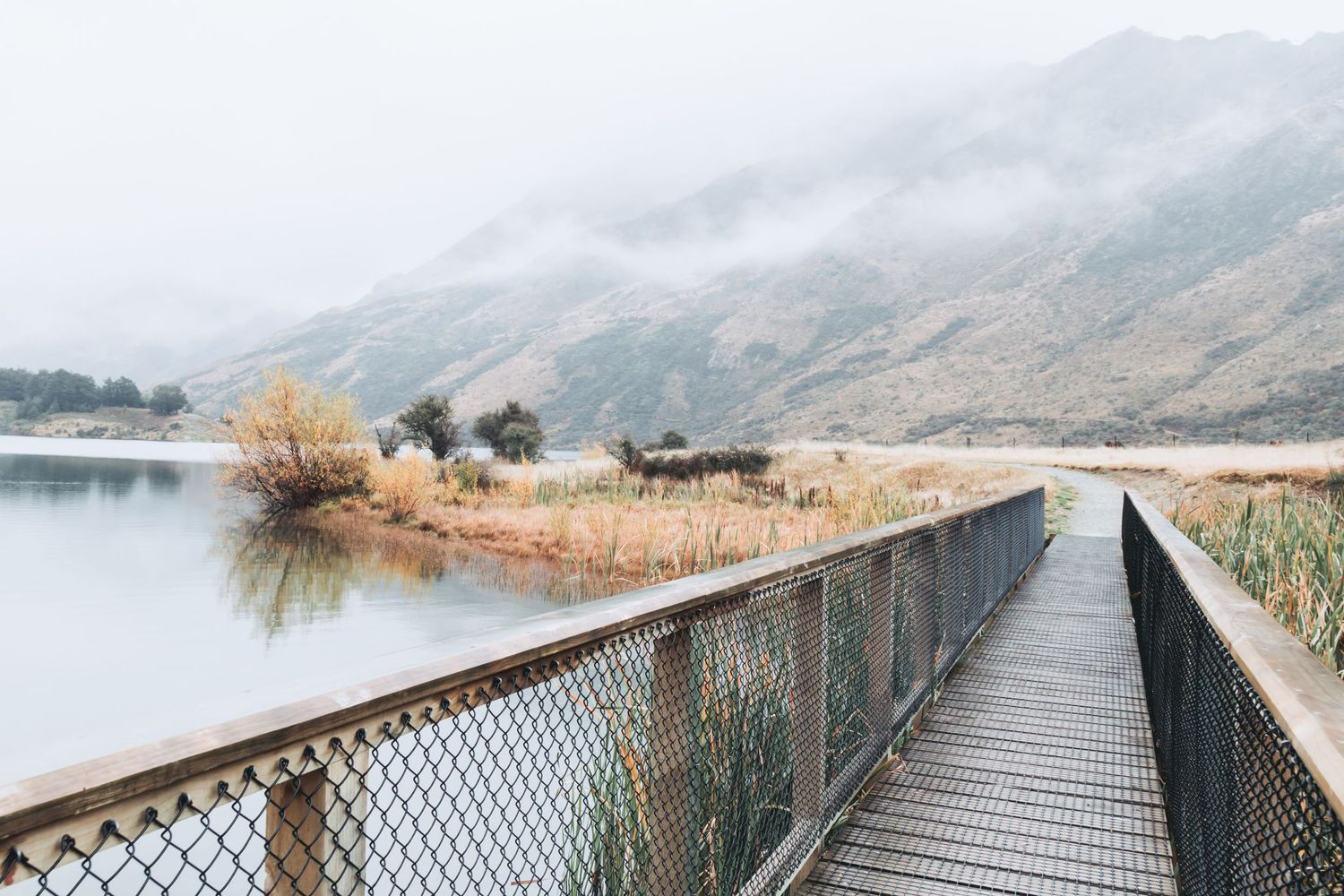 Wooden boardwalk extends over calm lake with misty mountains in the background.