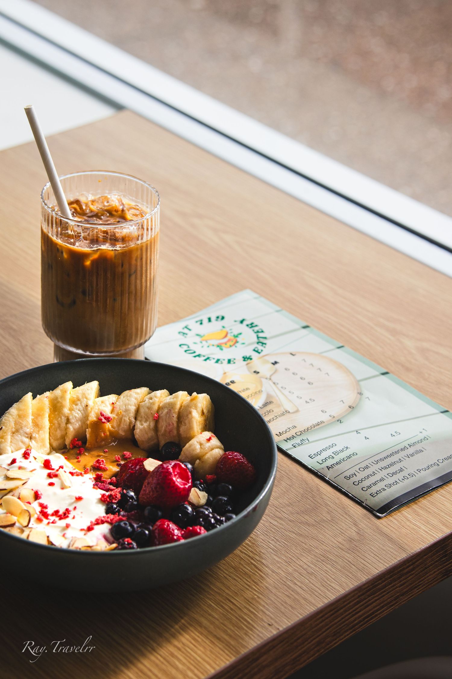 Breakfast bowl with sliced bananas and berries alongside iced coffee and menu on wooden table.