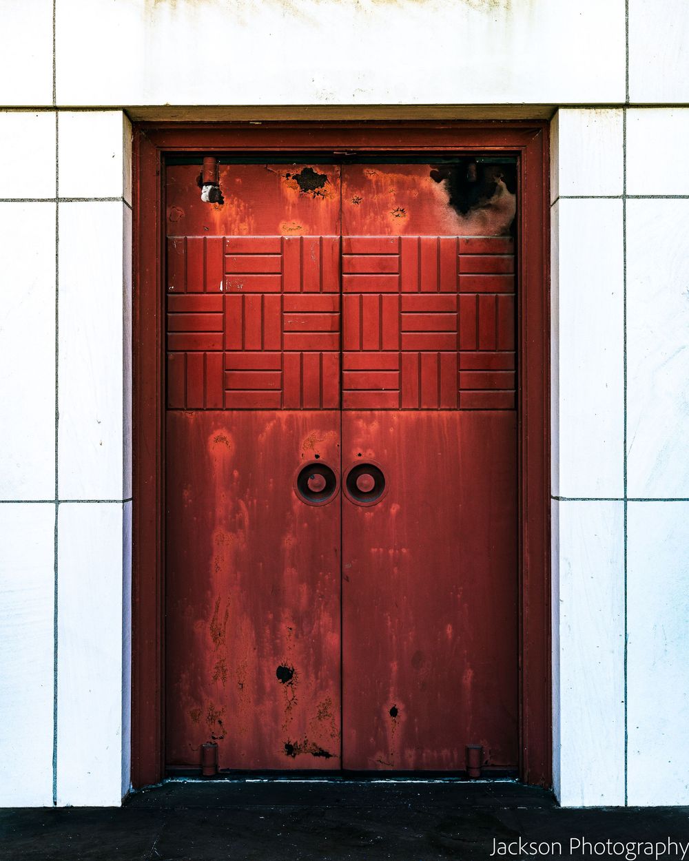 Weathered red double doors with geometric panel designs set in a white tiled wall, captured by Jackson Photography.