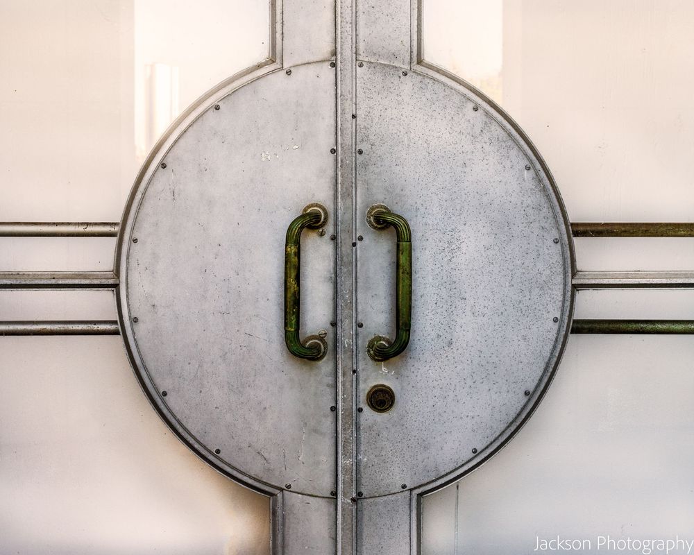 Art deco metal double doors with circular design and green patina handles on a light background.