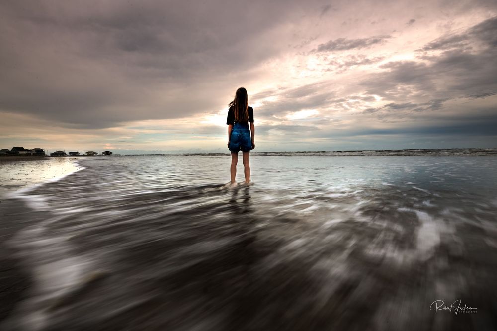 A lone woman stands barefoot on a reflective wet beach at sunset, gazing at the dramatic cloudy sky over the ocean.