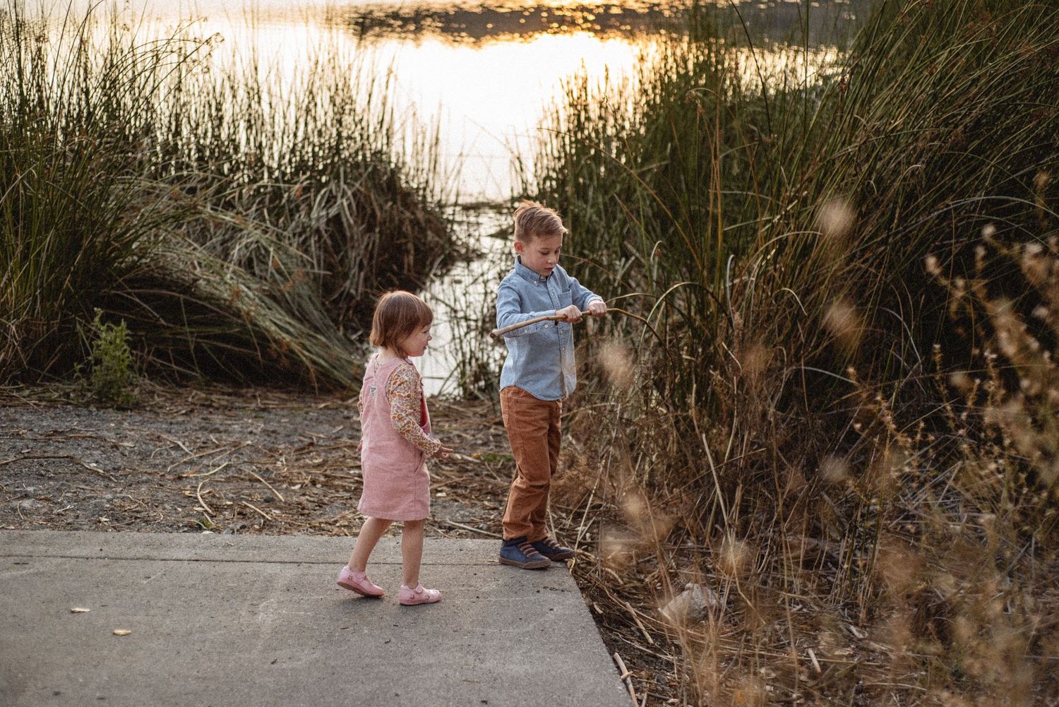 Two children standing on path near water during sunset.