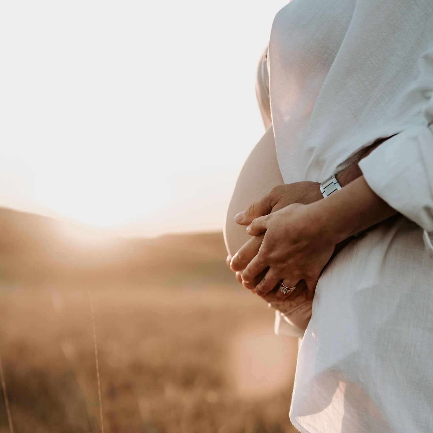 Close-up of pregnant woman cradling baby bump with partner's hands in warm sunset light outdoors.