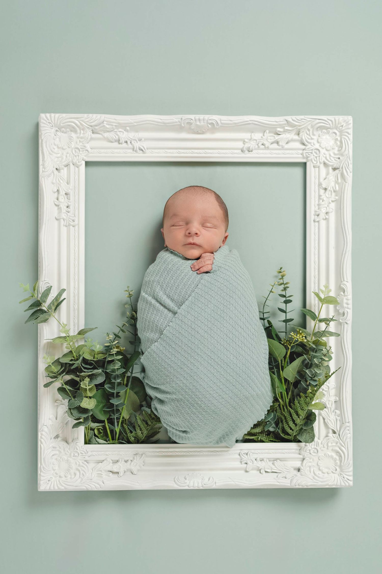 Sleeping baby wrapped in green swaddle inside white frame with eucalyptus leaves. Gosport newborn photography, fareham baby photographer