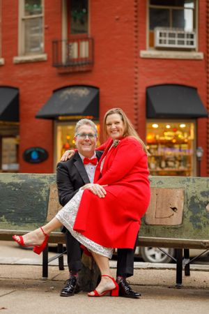 A romantic couple sits on a bench outside a brick building with black awnings, the woman wearing a striking red coat.