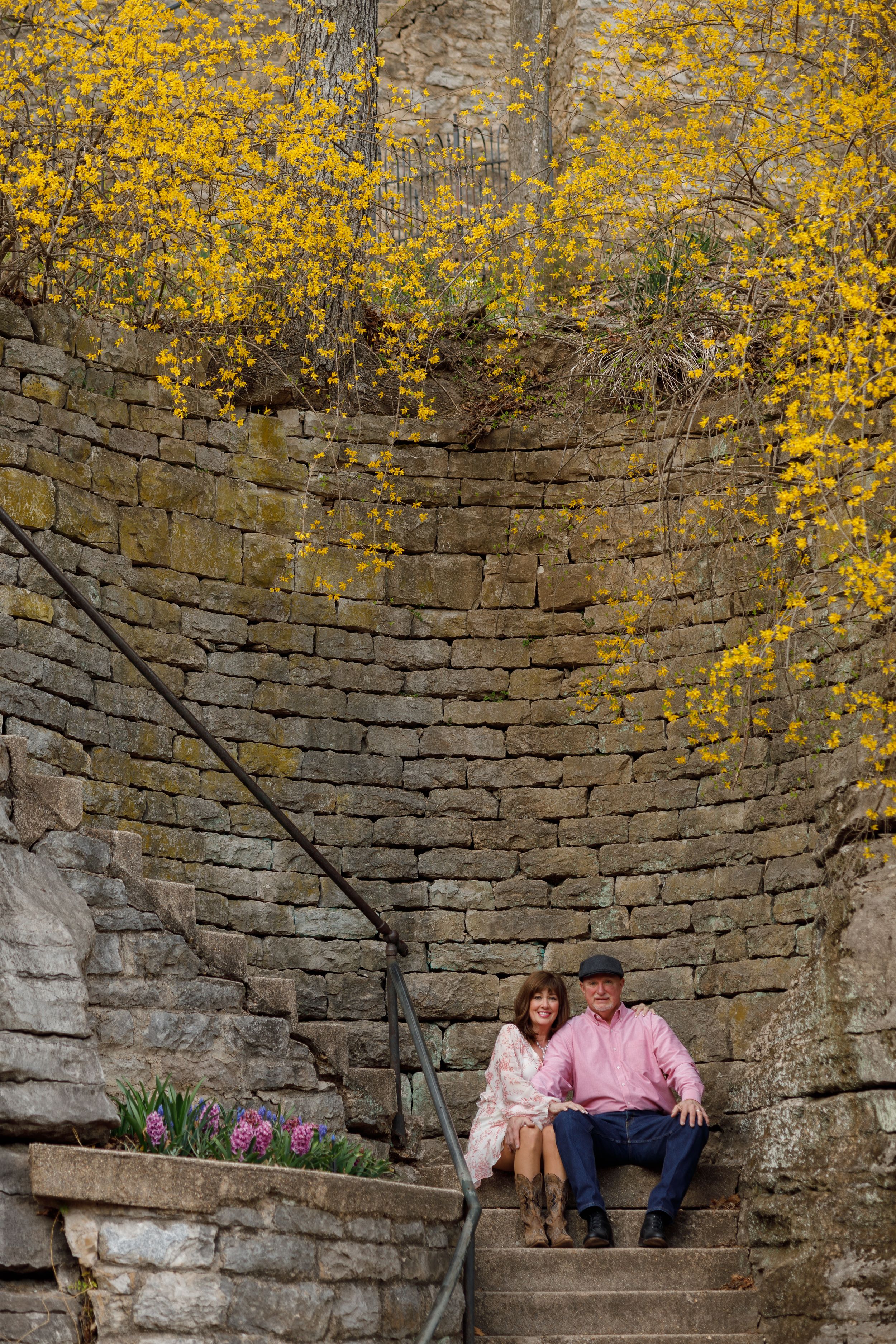 A couple sits on stone steps against an old brick wall with bright yellow forsythia blooming above in springtime.