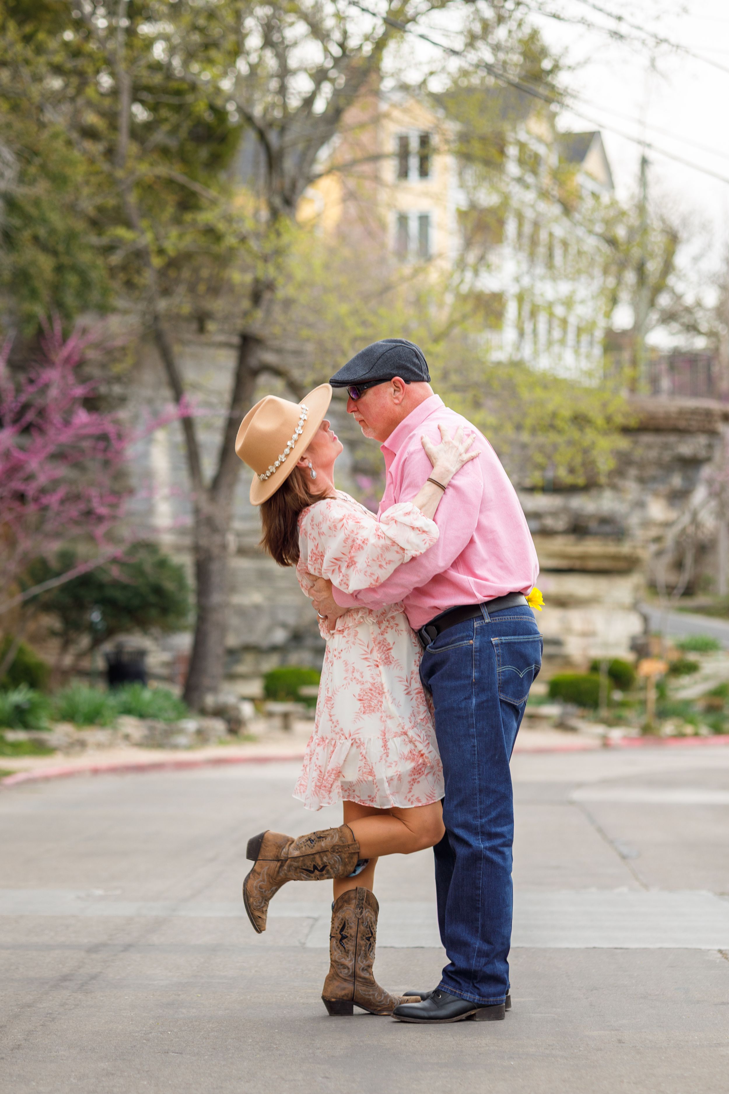 A couple embraces on a sidewalk in front of a garden, wearing cowboy boots and western-inspired outfits in springtime.