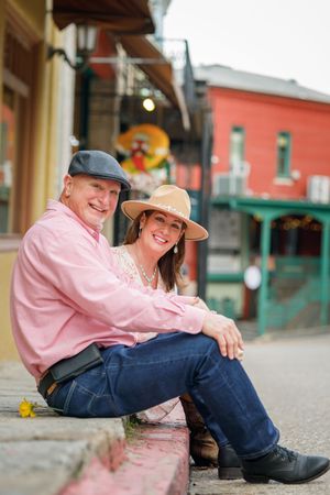 Couple sitting together on curb in casual attire with pink and red buildings in background on sunny day.