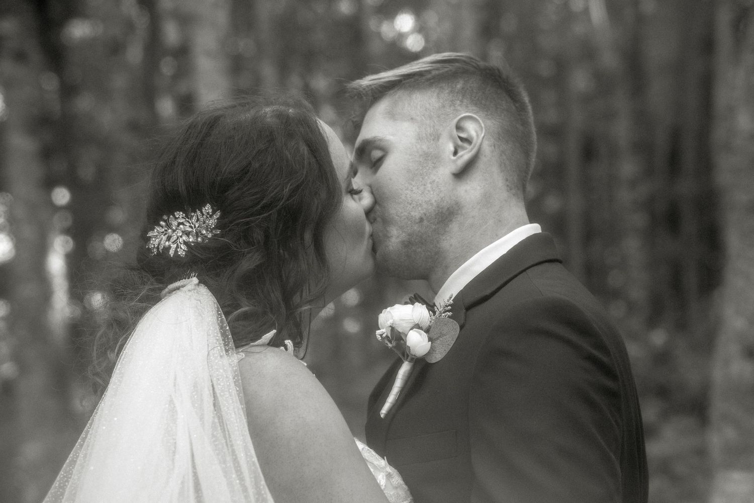A tender kiss shared between newlyweds in a forest setting, captured in romantic black and white photography. Photo at Clinton Hills