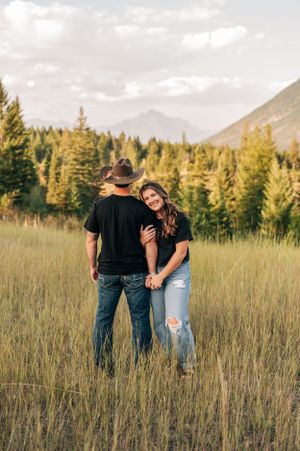 Couple holding hands while standing in mountain meadow with forest backdrop.
