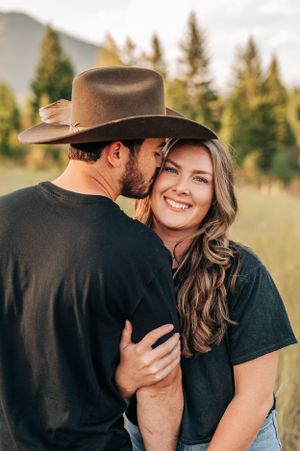 A romantic couple embraces in a field with mountains and evergreen trees in the background at golden hour.