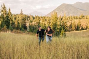 A couple walks through tall grass with mountains and forest behind them during golden hour.
