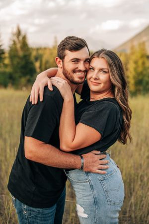 Couple in matching black shirts embrace and smile in a mountain meadow during golden hour.