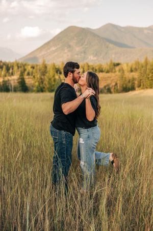Couple sharing a romantic kiss in a mountain meadow with evergreen forest backdrop.