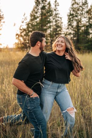 A couple laughs together in a golden field at sunset with evergreen trees in the background.