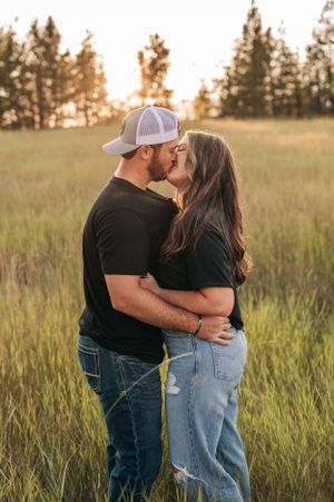 A romantic couple embraces in a sunlit meadow with pine trees silhouetted against the evening sky.