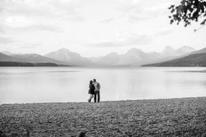 Two silhouettes stand together on a rocky shore with mountain peaks and a serene lake in the background.