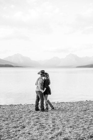 A romantic silhouette of a couple embracing on a lakeside beach with mountains in the distance.