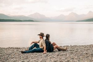 A couple sits back-to-back on a rocky beach overlooking a mountain lake at sunset.