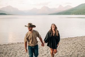 A couple walks hand in hand along a rocky beach with misty mountains across the lake.
