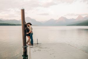 A couple embraces on a wooden pier with a scenic mountain lake backdrop.