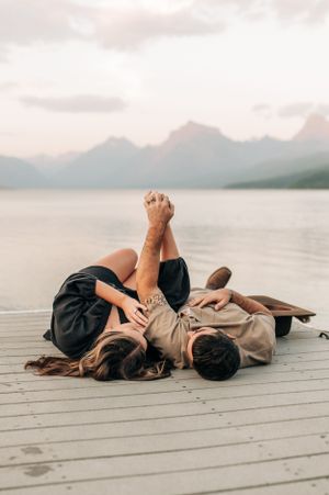 Couple lying on wooden dock by mountain lake at sunset sharing intimate moment together.