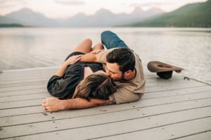 Romantic couple cuddling on lakeside dock with mountains in background during golden hour.
