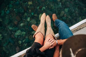 Overhead view of couple's feet dangling over water's edge with hands clasped together.
