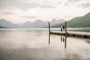 Silhouetted figures standing on wooden pier overlooking serene mountain lake at dusk.