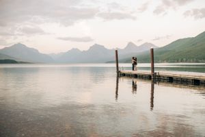 Lone figure reflected in calm mountain lake while standing on wooden dock at sunset.