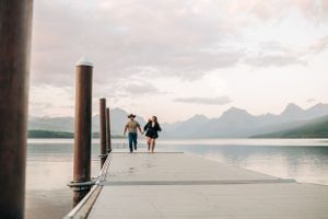 Two people walking down long wooden pier surrounded by mountain landscape and lake.